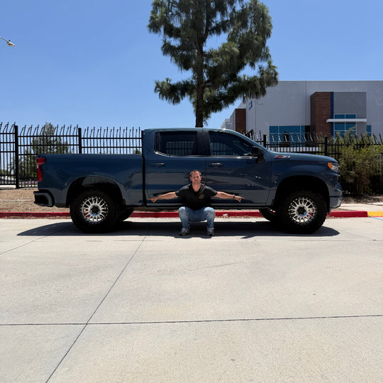 Man squatting with arms outstretched in front of a dark pickup truck, demonstrating tire clearance after mud flap delete installation