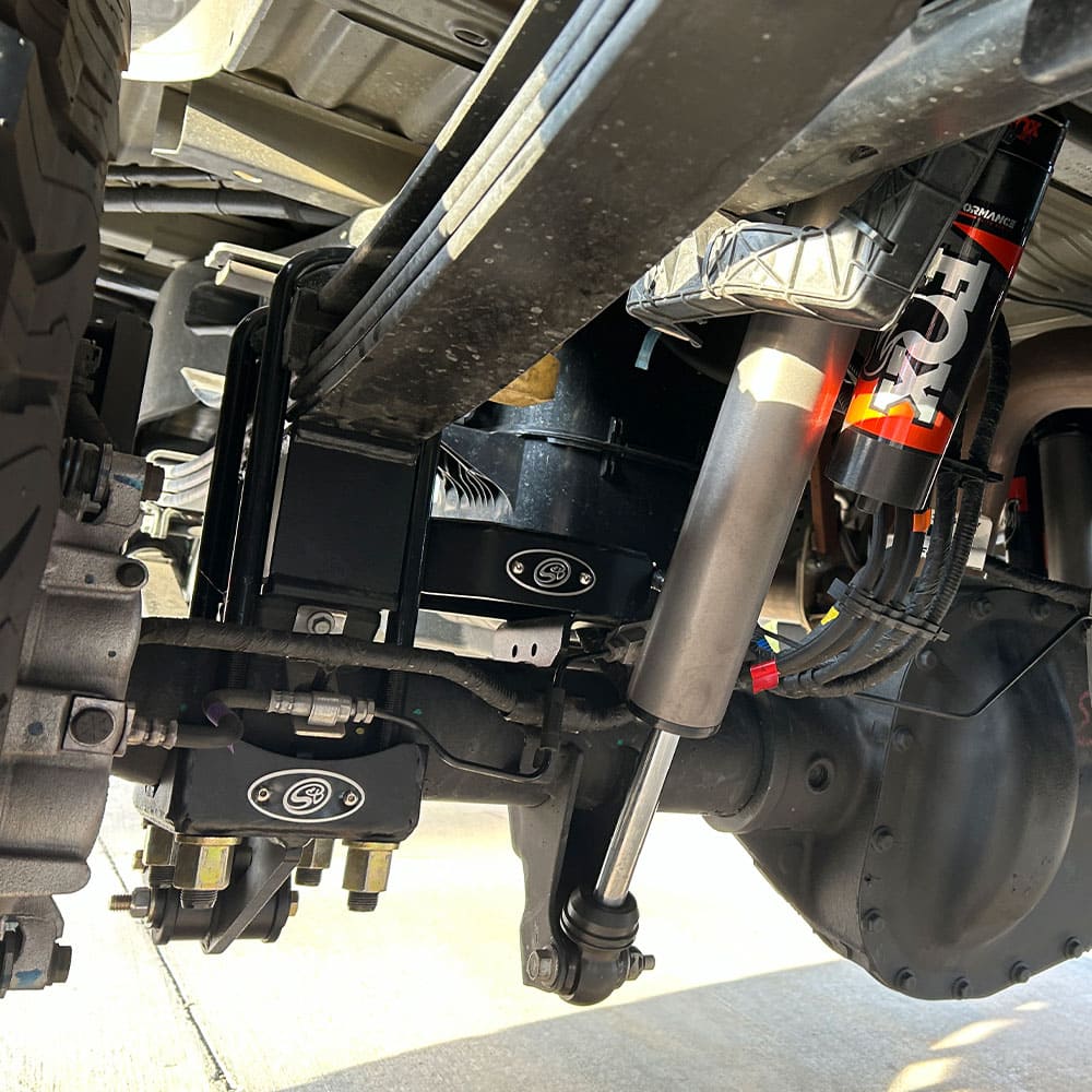 A S&B black 4" lift block installed on the lower control arm of a vehicle's suspension system.