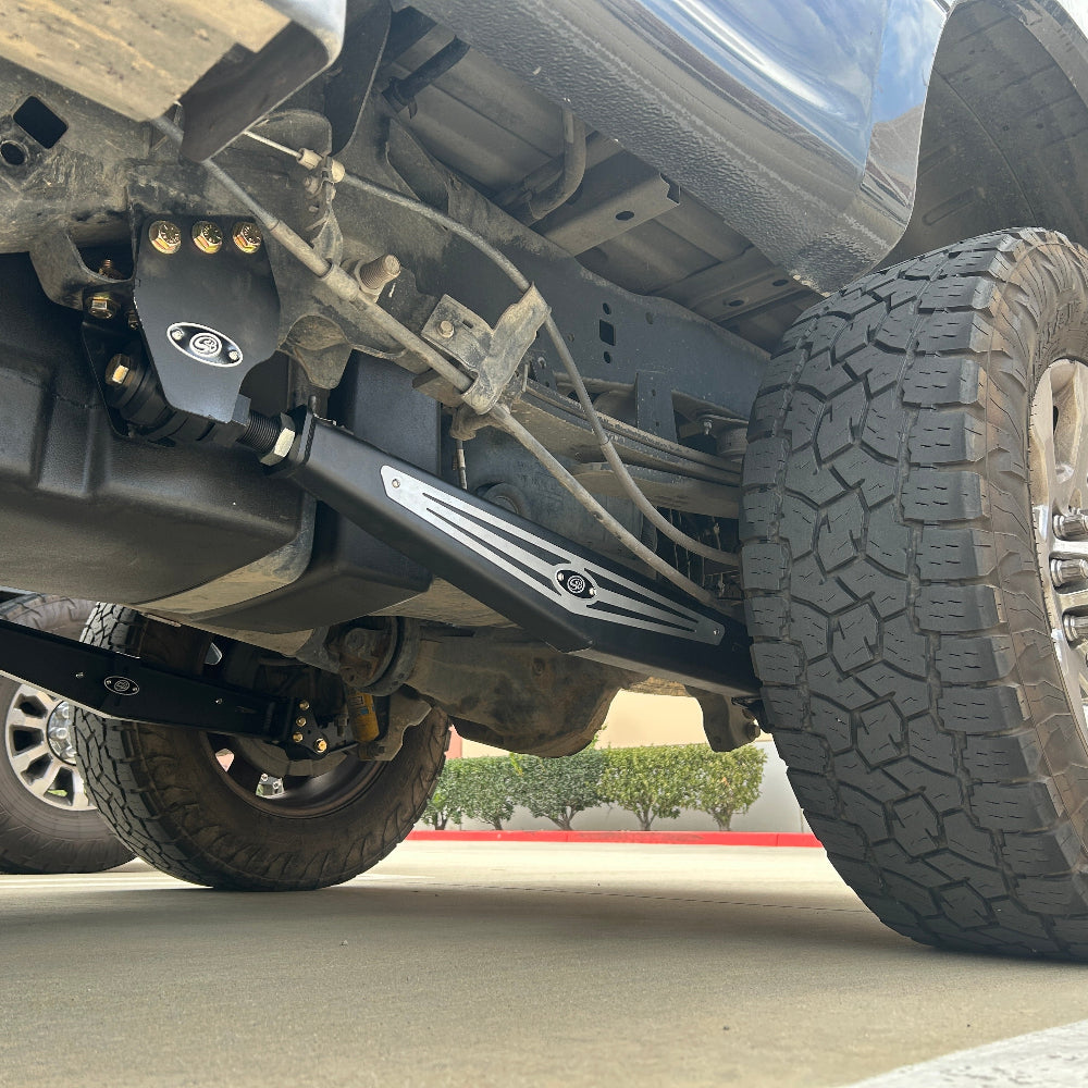 Close-up of a black metal undercarriage bar with silver decorative stripes and a circular logo, mounted near a large off-road tire on a vehicle.